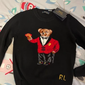 Svart stickad tröja Polo Ralph Lauren - Svart stickad tröja från Polo Ralph Lauren med ikonisk björn i smoking och röd kavaj på framsidan. RL-broderi i gult nere vid fållen. Rund halsringning och ribbade muddar. Perfekt för dig som gillar statement-plagg och klassisk preppy stil.
