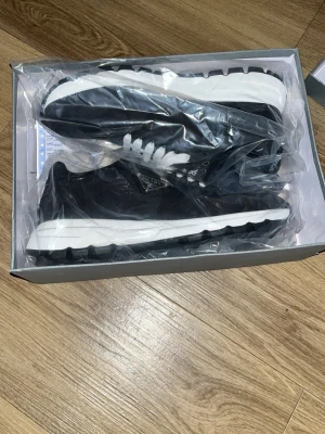Svarta Prada sneakers med vit sula - Stilrena svarta sneakers från Prada med vit, chunky sula och vita skosnören. Skorna har en klassisk låg modell och är tillverkade i textil med detaljer i läder. Sidan pryds av Pradas ikoniska metallogo. Perfekta för dig som vill ha en clean och lyxig look.