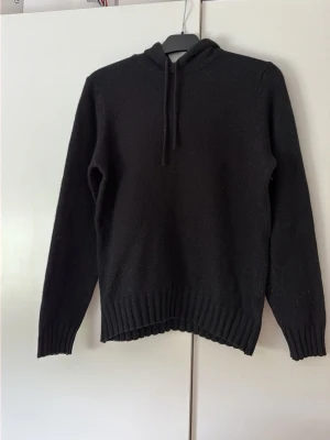 Svart stickad hoodie - Svart stickad hoodie med långa ärmar och ribbstickade muddar. Tröjan har huva med dragsko och är tillverkad i ett mjukt, värmande material. Perfekt för kyliga dagar när du vill vara både bekväm och snygg.