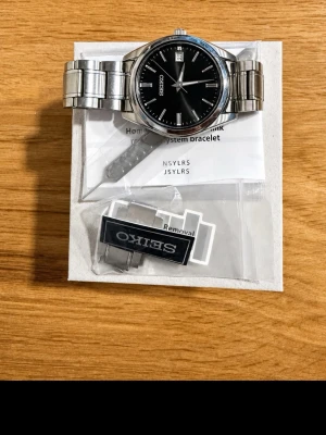 Seiko klocka med svart urtavla - Stilren Seiko klocka i rostfritt stål med svart urtavla och datumvisning. Klockan har en klassisk rund form och länkar i silverfärgat stål. Extra länkar och originaltillbehör medföljer. Perfekt accessoar för dig som gillar tidlös design.