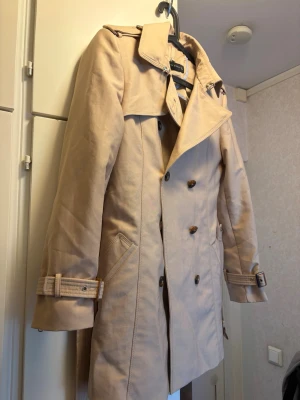 Beige trenchcoat från Zara Woman - Stilren beige trenchcoat från Zara Woman med dubbelknäppning, klassisk krage och bälte vid ärmsluten. Jackan har två snedställda fickor och axelklaffar för en tidlös look. Perfekt för dig som vill ha en snygg och trendig ytterjacka till vardagen.