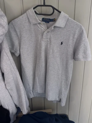 Grå pikétröja från Polo Ralph Lauren - Klassisk grå pikétröja från Polo Ralph Lauren i slim fit-modell. Tröjan har korta ärmar, krage och knäppning framtill samt den ikoniska broderade loggan på bröstet. Perfekt för en stilren och avslappnad look.