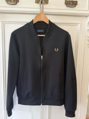 Fredd Perry svart zip hoodie  - Stilren svart bomberjacka från Fred Perry med guldfärgad dragkedja och broderad logga på bröstet. Jackan har ribbade muddar, två fickor framtill och klassisk bomberkrage. Materialet känns mjukt och bekvämt, perfekt för en clean och sportig look.