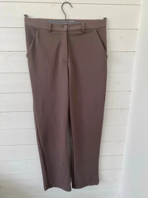 Bruna raka kostymbyxor - Snygga bruna kostymbyxor med raka ben och pressveck. Byxorna har hällor för skärp och stängs med knapp och dragkedja samt att de har fickor. Perfekta för en stilren look. Materialet känns mjukt och följsamt.