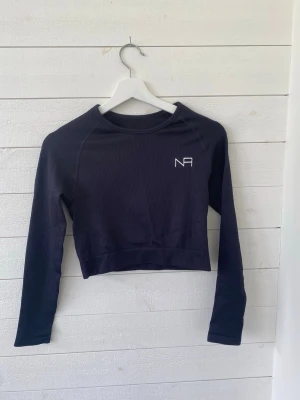 Svart croppad långärmad träningstopp NA - Svart croppad långärmad tränings topp från NA med enkel design och liten vit logga på bröstet. Perfekt för en sportig och trendig look. Mjuk och stretchig känsla, sitter tight mot kroppen.