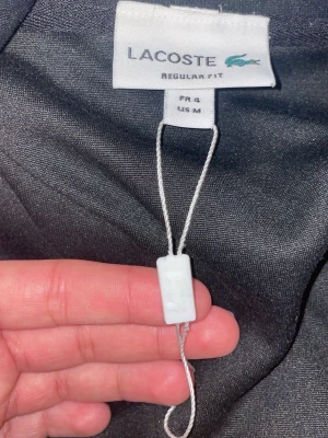 Svart hoodie från Lacoste - Snygg svart hoodie från Lacoste i regular fit. Klassisk design med huva och ribbade muddar. Tillverkad i mjukt material som känns skönt mot huden. Perfekt för dig som gillar stilrena och tidlösa plagg med en sportig touch.