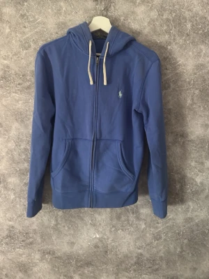Zip hoodie Polo Ralph Lauren - Snygg blå hoodie med dragkedja från Polo Ralph Lauren. Klassisk design med vit logga broderad på bröstet, huva med vita snören och två stora fickor framtill. Perfekt för en chill och avslappnad stil.