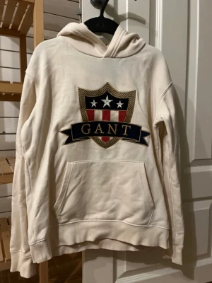 Vit hoodie GANT Med två fläckar på!  - Snygg vit hoodie från GANT med stor broderad sköldlogga i rött, blått och guld på bröstet. Klassisk känguruficka framtill och ribbade muddar. Perfekt för dig som gillar stilren och sportig look.                                      Hoodien har två fläckar! 