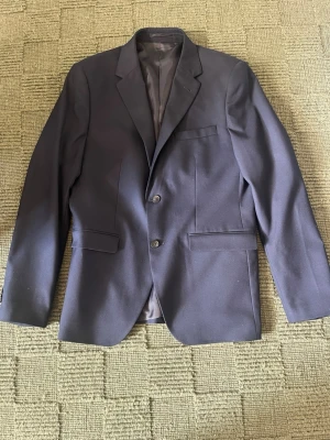 Mörkblå kavaj med två knappar - Slimfit mörkblå kavaj från Riley med två knappar. Kavajen har slät finish och är helfodrad på insidan. Det finns även byxor till som jag kan skicka bilder på om någon vore intresserad!
