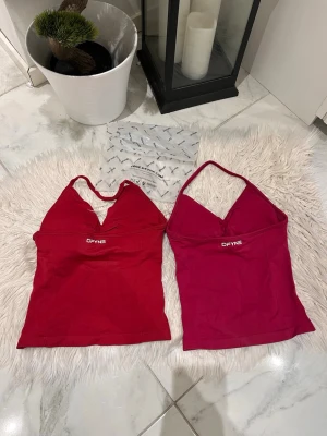 DFYNE äldre modell 2-pack - Säljer två snygga linnetoppar från DFYNE, en i röd och en i rosa. Båda har smala axelband, v-ringad hals och logga framtill. Perfekta för sommaren och tillverkade i mjuk, stretchig syntet. Snygg passform och trendig look! Modell: impact 