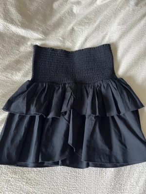 Marinblå volangkjol med smock - Supersnygg marinblå kortkjol med bred smock i midjan och dubbla volanglager. Kjolen har en fin passform och är perfekt för dig som gillar en trendig och bekväm stil. Tillverkad i mjukt tyg som känns skönt mot huden. Storlek 34