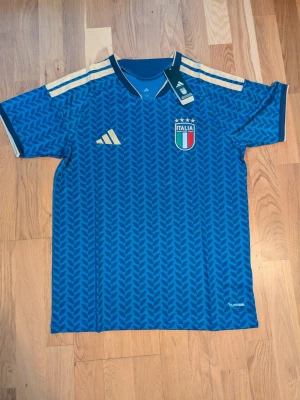 Italien Adidas fotbollströja blå. M - Helt ny Blå fotbollströja från Adidas med Italiens landslagsmärke på bröstet och guldiga detaljer på axlarna. Tröjan har ett mönster i blå nyanser, korta ärmar och Climalite-material som andas. Perfekt för träning eller match!