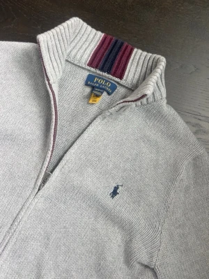 Grå ziptröja från Polo Ralph Lauren - Snygg grå ziptröja från Polo Ralph Lauren med broderad logga på bröstet. Tröjan har hög krage med ribbade detaljer och ränder i vinrött, blått och svart. Perfekt för lager-på-lager och har en klassisk, stilren look.