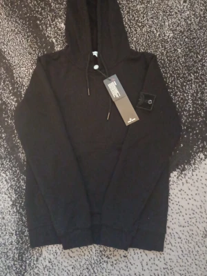 Svart hoodie från Stone Island - Snygg svart hoodie från Stone Island med klassisk huva och dragsko. Hoodien har en diskret patch på ärmen och ribbade muddar vid ärmslut och nederkant. Perfekt för dig som gillar streetwear och vill ha en stilren look.