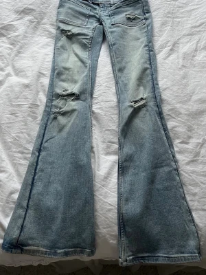 Ljusblåa lowwaist utsvängda jeans med hål - Säljer ett par ljusblåa jeans med utsvängda ben och slitningar på framsidan. Jeansen har klassiska fem fickor och en snygg tvättad look. Har en liten fläck på ena låret (se bild 4) men absolut ingenting som syns vid användning!