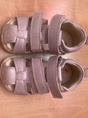 Ljusrosa sandaler med kardborreband storlek 23 - Säljer ett par ljusrosa sandaler med två breda kardborreband och öppen tå. Sandalerna har en vit sula med bra grepp och är tillverkade i mjukt läderliknande material. Insidan är ljus med diskret mönster och fjärilsdetalj.