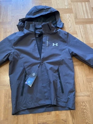 Mörkgrå vindjacka från Under Armour - Snygg mörkgrå vindjacka från Under Armour med huva, dragkedja och två framfickor med svarta dragkedjor. Jackan har logga på bröstet och meshfoder i huvan. Perfekt för blåsiga dagar och sportiga aktiviteter. Den passar storleke S och förmodligen M. Aldrig använt den