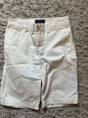 Vita shorts från Polo Ralph Lauren - Klassiska vita shorts från Polo Ralph Lauren med normal passform och midwaist. Tillverkade i mjuk bomull med bälteshällor, knappgylf och två fickor fram samt två bakfickor med knapp. Perfekta för sommardagar. Storlek 140.