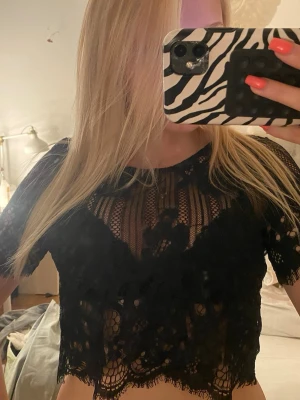 Svart spetsig croppad topp - Snygg svart croppad topp i spets med korta ärmar och transparenta detaljer. Toppen har ett feminint mönster och vågig kant nertill, perfekt för att lyfta vilken outfit som helst.