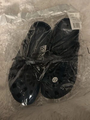 Mörkblå tofflor i plast, storlek 37 - Säljer ett par mörkblå tofflor i plast med ventilerande hål och enkel slip-in design. Perfekta att ha hemma eller på stranden. Tofflorna är lätta och smidiga att ta på sig. Märke och ursprungsland syns på etiketten.
