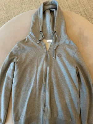Grå zip-hoodie från Knowledge Cotton Apparel - Grå hoodie med dragkedja från Knowledge Cotton Apparel. Klassisk modell med huva och dragsko, samt rund logotyp-patch på bröstet. Tillverkad i mjuk bomullsblandning, perfekt för en avslappnad stil.