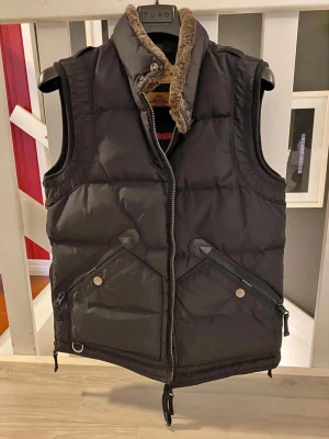 Svart dunväst från Parajumpers - Stilren svart dunväst från Parajumpers med hög krage och mjukt foder. Västen har två stora fickor framtill med dragkedja och tryckknappar samt detaljer i orange på sidorna. Perfekt för lager-på-lager och snygg till streetwear.