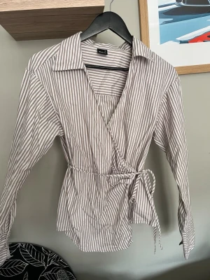 Randig omlottblus med knytband från Gina  - Säljer en beige och vit randig omlottblus från ginatricot. Blusen har långa ärmar, klassisk krage och knytband i midjan för en snygg passform. Tillverkad i ett lätt och bekvämt tyg, perfekt för en trendig och avslappnad look. Aldrig använd men köpt i höstas💕
