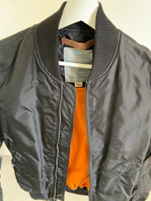 Svart bomberjacka från Alpha Industries - Klassisk svart bomberjacka från Alpha Industries med orange foder och ribbade muddar. Jackan har dragkedja framtill, fickor på sidorna och en ärmficka med röd tag. Perfekt för en cool streetstil.