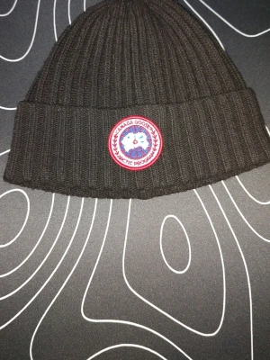 Svart ribbstickad mössa Canada Goose - Säljer en svart ribbstickad mössa från Canada Goose med klassisk uppvikt kant och det ikoniska runda röda och vita märket framtill. Mössan är tillverkad i ett mjukt och varmt material, perfekt för kalla dagar.