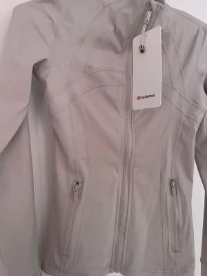 Lululemon set - Snygg ljusgrå jacka från Lululemon med figurnära passform. Jackan har dragkedja framtill, två fickor med dragkedja och diskreta sömdetaljer som ger en sportig look. Perfekt för dig som gillar stilrena träningskläder.