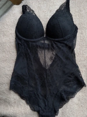 Svart spetsbody med vadderade kupor - Snygg svart body i genomskinlig spets med vadderade kupor och bygel. Djup urringning och fina spetsdetaljer längs kanterna. Perfekt för att känna sig extra fin. Storlek 75B i kupa och bodyn är i XS🥰