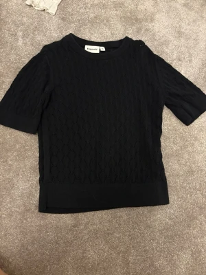 Svart stickad topp från Kappahl - Snygg svart stickad topp från Kappahl med korta ärmar och diskret mönster i tyget. Rund halsringning och ribbade kanter ger en clean look. Perfekt att matcha med både jeans och kjol.