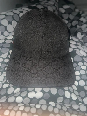 Gucci keps svart canvas  - Snygg svart keps från Gucci med klassiskt GG-monogram över hela kepsen. Justerbar rem baktill och böjd skärm. Tillverkad i canvasmaterial som ger en exklusiv känsla och passar perfekt till streetwear eller en trendig look. OBS SÄLLSYNT I DETTA SKICK DÅ DET ÄR GAMLA MODELLEN