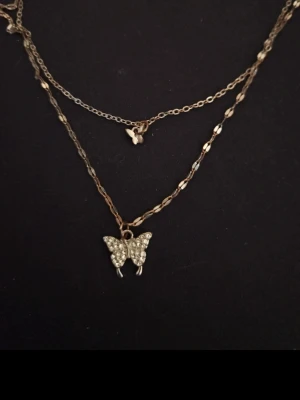 Silvrigt halsband med fjärilar - Dubbelkedjat halsband i silver med två fjärilshängen. Kedjorna har olika länkar och ger en trendig lager-på-lager-look. Perfekt accessoar för att lyfta din outfit.