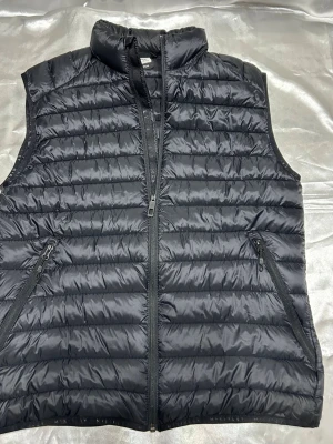 Svart dunväst från McKinley - Snygg svart dunväst från McKinley med quiltad design och hög krage. Västen har två dragkedjeförsedda fickor på sidorna och stängs med dragkedja framtill. Perfekt för lager-på-lager och håller dig varm under kyliga dagar.