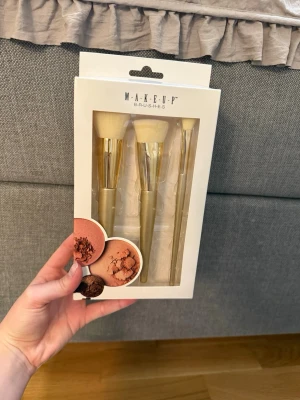 Make-up borstar set i guld - Snyggt set med tre sminkborstar från Make-Up Brushes. Borstarna har guldfärgade handtag och ljusa, mjuka borst. Perfekta för applicering av puder, rouge och highlighter. Kommer i en stilren förpackning med makeup-tema.