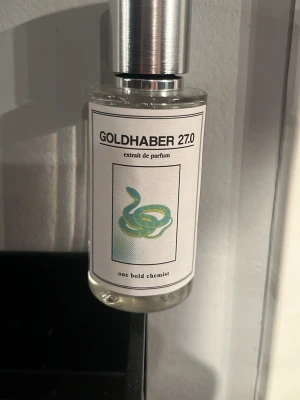 Goldhaber 27.0 Parfym One Bold Chemist - Säljer en stilren parfymflaska från one bold chemist, Goldhaber 27.0. Flaskan är genomskinlig med en silverfärgad metallkork och har en vit etikett med en grön och blå orm som motiv. Modern och minimalistisk design, perfekt för dig som gillar unika dofter.