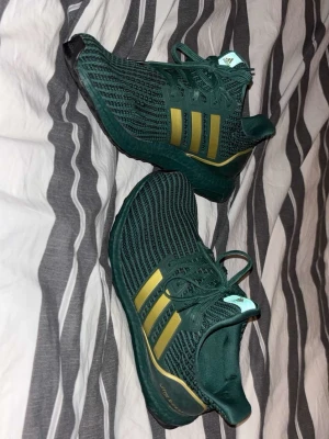 Adidas Ultraboost gröna/guldiga  - Snygga Adidas Ultraboost sneakers i mörkgrönt med guldiga ränder på sidorna. Ovandelen är i stickad textil för extra komfort och sulan är svart med vit detalj undertill. Perfekta för dig som gillar sportig och modern stil.