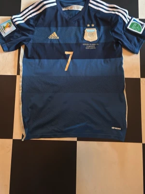 Argentina Di Maria matchtröja blå - Säljer en blå Argentina fotbollströja från Adidas med nummer 7 och Di Maria på ryggen. Tröjan har guldiga detaljer, två stjärnor ovanför landslagsemblemet och officiella VM-märken på ärmarna. Tillverkad i lätt funktionsmaterial (climacool).