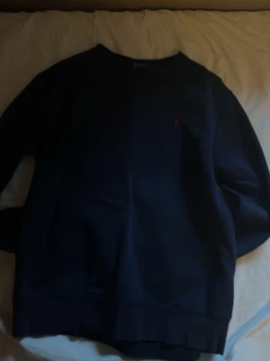 Mörkblå crewneck från Polo Ralph Lauren - Mörkblå sweatshirt med klassisk crewneck från Polo Ralph Lauren. Tröjan har en broderad röd logga på bröstet och är tillverkad i mjukt bomullsmaterial. Perfekt för en avslappnad och stilren look.