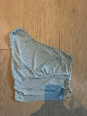 Ljusblå bikiniöverdel med en axel - Trendig ljusblå bikiniöverdel med asymmetrisk design och en axel. Tillverkad i ett mjukt, ribbat material som ger en snygg och bekväm passform. Perfekt för strandhäng eller poolparty.