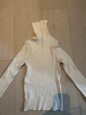 Beige ribbstickad polotröja - Snygg beige ribbstickad polotröja med lång ärm och hög krage. Tröjan är figurnära och har en mjuk, stretchig känsla. Perfekt att styla med jeans eller kjol för en trendig look.