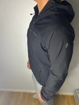 Svart vindjacka från peak performance  - Snygg svart vindjacka från peak performance med huva och dragkedja framtill. Jackan har två sidofickor med dragkedja och diskret logga på bröstet samt ärmen. Tillverkad i ett lätt och smidigt material som passar perfekt för blåsiga dagar.🙌