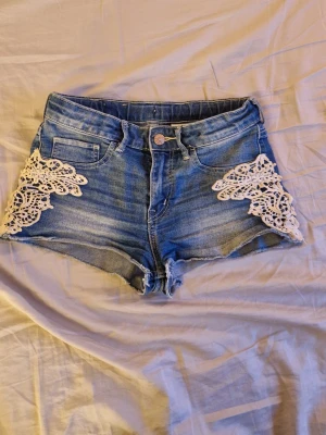 Blå jeansshorts med spetsdetaljer - Snygga blå jeansshorts med hög midja och vita spetsdetaljer på sidorna. Klassisk femficksmodell med råa kanter nertill och knappstängning framtill. Perfekta för varma dagar och en trendig look.