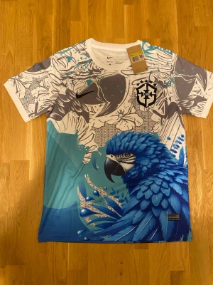 Nike Brasil CBF t-shirt med blå papegoja - Unik sporttröja från Nike och Brasilien CBF med färgstarkt tryck av en blå papegoja framtill. Tröjan har vita och grå partier med grafiska mönster, rund hals och korta ärmar. Materialet är lätt och andas, perfekt för träning eller sport.