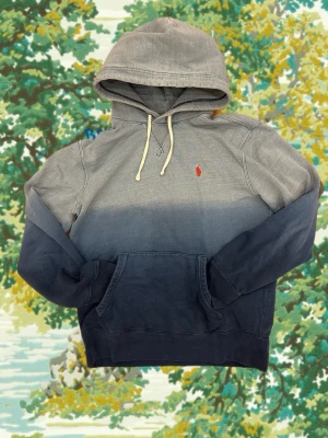 Blå hoodie från Polo Ralph Lauren - Snygg hoodie från Polo Ralph Lauren i en cool blå ombre-färg. Klassisk huva med dragsko och stor magficka framtill. Liten röd logga broderad på bröstet. Perfekt för en avslappnad och trendig look.