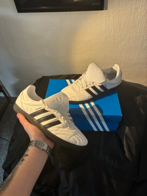 Adidas Samba OG vita sneakers - Adidas Samba OG sneakers i vitt läder med svarta detaljer och klassiska tre ränder på sidorna. Yttersulan är svart och skon har en låg profil. Tungan är extra lång och pryds av Adidas-loggan. Perfekt för dig som gillar retrostil och streetwear.