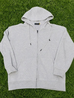 Grå zip-hoodie från Polo Ralph Lauren - Snygg grå hoodie med dragkedja från Polo Ralph Lauren. Klassisk design med huva, dragsnören och broderad logga på bröstet. Mjuk bomullsblandning och två fickor framtill. Perfekt för en chill och stilren look.