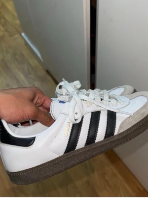 Adidas Samba OG, 41 1/3 - Adidas Samba OG sneakers med vit ovandel och svarta ränder. Skorna har en gråbrun yttersula och snörning framtill. ”SAMBA” är tryckt i guld på , ny pris 1300kr, storlek känns som 41-42. Säljer då jag vill bli av med dessa och behöver en ny ägare