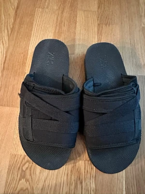 Svarta tofflor från ZARA med kardborre - Säljer ett par svarta tofflor från ZARA med breda kardborreband över foten. De har en sportig och modern design med textilöverdrag och mönstrad sula för extra grepp. Perfekta för att chilla hemma eller snabbt dra på sig när man ska ut.